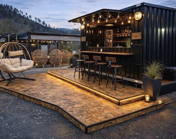 Black container bar at dusk with a lit wood deck, bar stools and lounge chairs